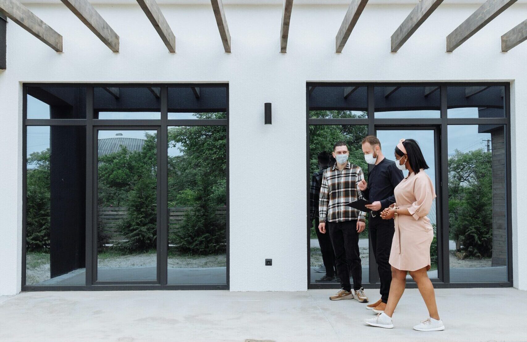 Three real estate agents wearing masks showcase a sleek, modern home exterior to clients.